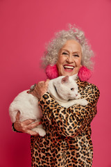 Pleased fashionable elderly woman holds domestic pet, feels not lonely in company of cat, dressed in fashionable leopard shirt, smiles broadly, going to have walk with animal, relax together at home