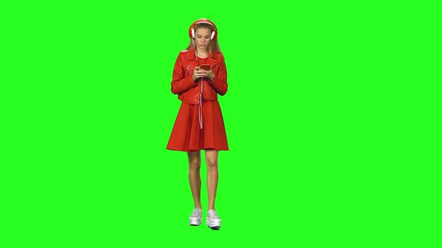 Blonde girl is walking in big red headphones, chooses music and moves to rhythm at green screen