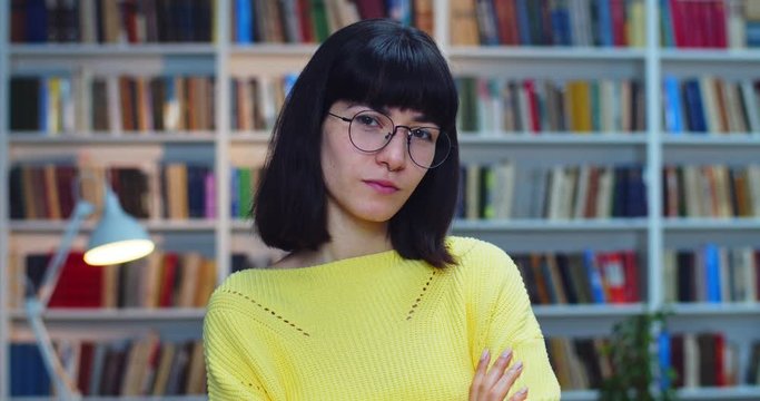 Close-up portrait of a serious pretty woman with braces blach short hair and yellow sweater standing close to bookshelf in the library and crossing her arms.