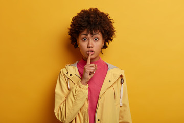 Surprised ethnic woman tells secret and asks keep quiet, presses index finger to lips, promises stay silent, has impressed expression, hides secret and gossips, wears casual anorak, isolated on yellow