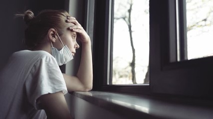 slow motion side view closeup of worried young Caucasian woman with blonde hair and white shirt wearing medical protection face mask watching the city by the window quarantine concept