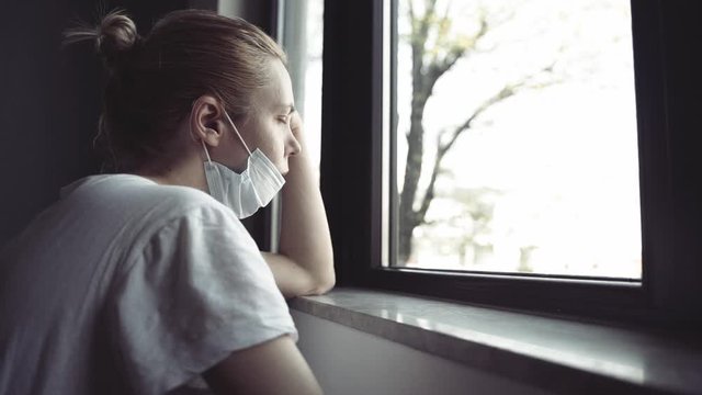 slow motion side view closeup of worried young Caucasian woman with blonde hair and white shirt wearing medical protection face mask watching the city by the window quarantine concept