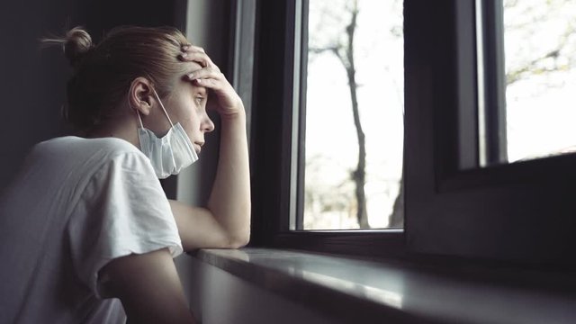 slow motion side view closeup of worried young Caucasian woman with blonde hair and white shirt wearing medical protection face mask watching the city by the window quarantine concept
