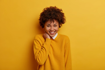 Fototapeta na wymiar Beautiful optimistic woman with smooth skin and white teeth smiles tenderly, touches neck, raises mood as watches comedy at home, wears bright yellow jumper, sends good vibes, models in studio