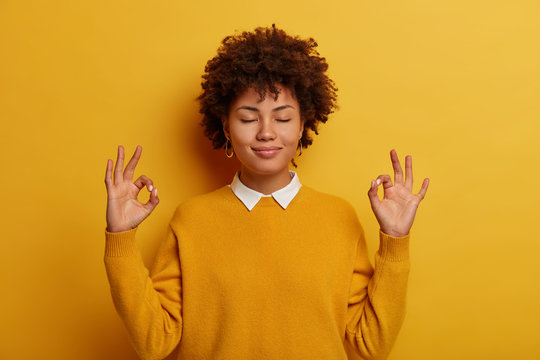 Young woman with closed eyes, makes yoga gesture, meditates to feel relieved and stress free, has patient calm expression, breathes deeply, stands in lotus pose, isolated on yellow background