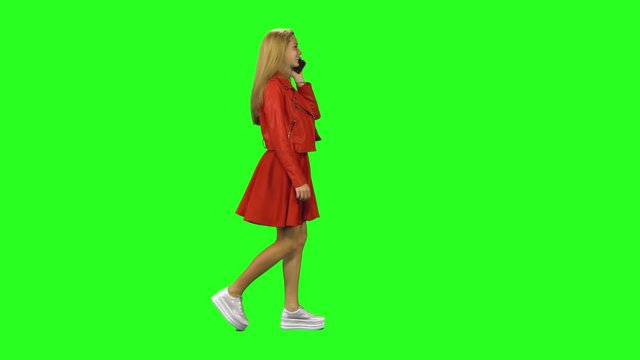 Blonde girl is calmly walking and talking on the mobile phone at green screen background. Chroma key. Profile view.