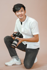Asian man in white shirt kneeling while holding digital camera in hands. Male photographer looking...