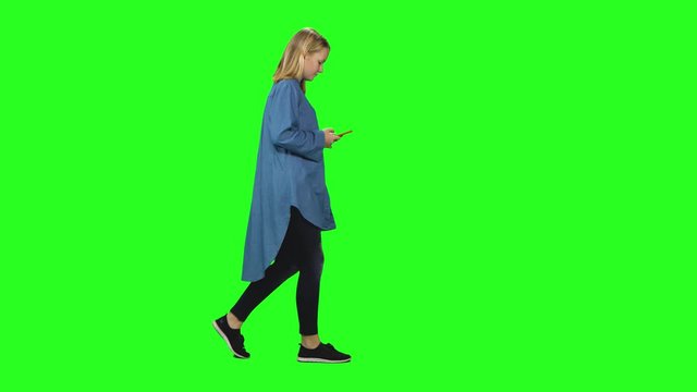 Blonde teenager girl calmly walking and reading text message on her mobile phone at green screen. Profile view.