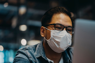 Asian young man indoors in cafe. Lifestyle concept photo with copy space. Picture with handsome guy who using protective face mask. Portrait with gray laptop