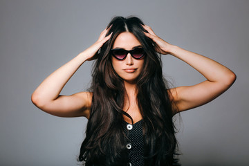 Beautiful young woman in sunglasses looking at camera with hands near her face on grey background.