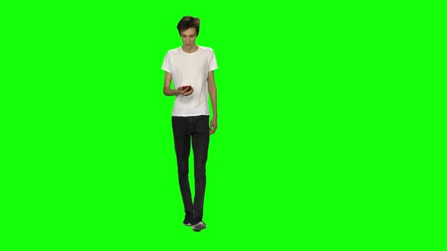 Tall skinny teen guy calmly walking and texting message vie his mobile phone on green screen. Chroma key. Front view.