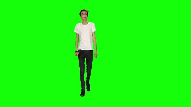 Tall skinny teen guy calmly walking and drinking coffee on green screen background. Chroma key, 4k shot. Front view.