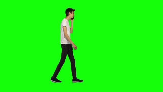 Tall skinny teen guy calmly walking and talking on the mobile phone on green screen. Chroma key, 4k shot. Profile view.