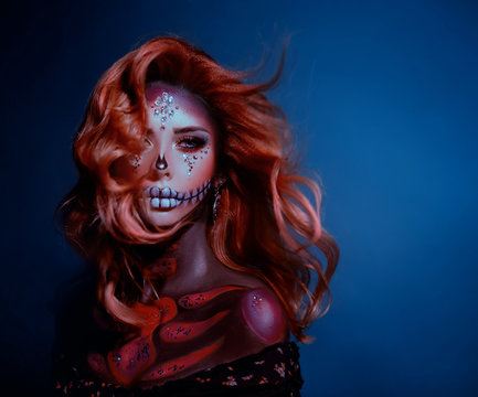 Artistic portrait young woman image sugar skull. Long redhaired wavy hair flies in wind. Stage creative makeup decorated with bright shiny sparkling crystal rhinestones. Backdrop blue color. Halloween