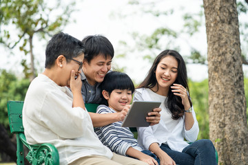 Asian happy family play in park, read electronic book from tablet together. Boy sit in on bench between grandmother and mother. Father hug him from back, point interest thing. Family lifestyle concept