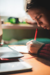 Young caucasian girl in red sweater with confused look on her face writing in notebook while studying online at home with pen in hand