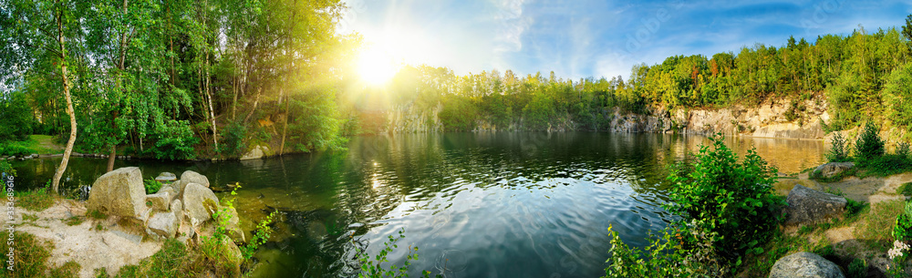 Wall mural panoramic landscape shot of idyllic lake surrounded by trees and cliffs, with the sun glowing on the