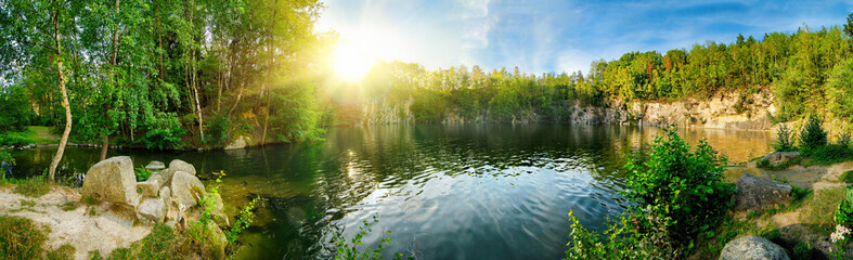 Panoramic landscape shot of idyllic lake surrounded by trees and cliffs, with the sun glowing on the horizon