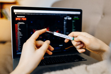 Electronic thermometer in a female hands. Woman lying on couch with laptop, measuring body temperature. Сoronavirus disease statistics on blurred screen background.