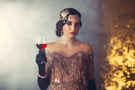 Closeup portrait flapper woman with glass wine. holiday headband finger wave hairstyle, vintage style 20s dress evening makeup red lips smokey cat eyes. Backdrop golden room smoke. Elegant prom image