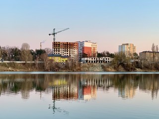 The construction of a high-rise building on the shore of the reservoir. A new building and a high-rise crane are reflected in the water of the lake. Apartments in a new house overlooking the river.