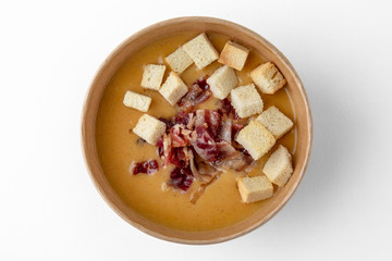 Tasty healthy hot pumpkin, lentil, pea, or potato cream soup with slices of bacon and crackers in a cardboard bowl  for take away or food delivery isolated on a white background.