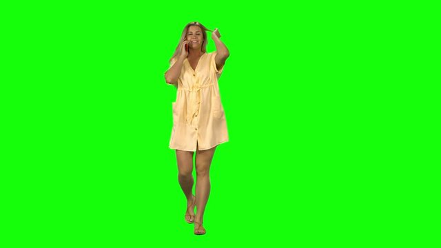 Tanned blond woman is calmly walking and talking on the mobile phone with smile on green screen. Front view.