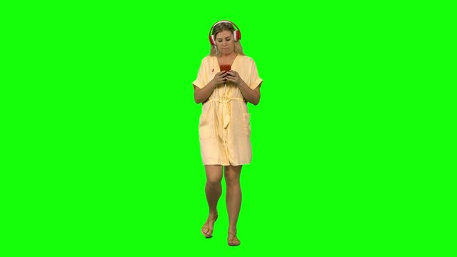 Tanned blond woman is calmly walking in big red headphones, chooses music on the mobile phone on greenscreen. Front view
