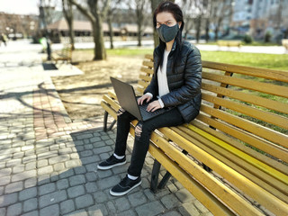 The masked girl is sitting on a bench in the park with a laptop and looking for work remotely during quarantine. Self-isolation and work remotely