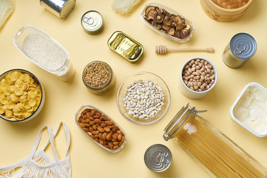 Creative flatlay with pantry staples. Glass jars with pasta, beans and chickpeas, canned goods, nuts and dried mushrooms. Top view pattern with basic products on yellow background