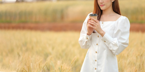 Fototapeta na wymiar Asian young woman holding hot paper cup coffee in barley field happiy, relaxing lifestyle, city break in nature, young traveler on vacation 