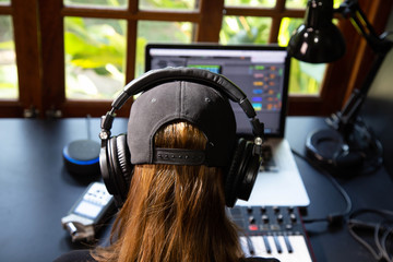Close up of a female music producer with headphones in her home studio, desk with digital recorder, Smart Home Assistant, notebook and a Midi keyboard. Window with nature in the background.