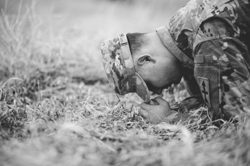Grayscale shot of an American soldier kneeling and praying on a dry grass field