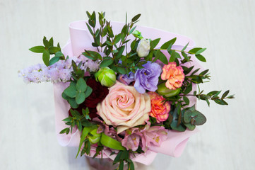 A delicate bouquet of different flowers in a light pink wrapper stands on a light background in a home setting.