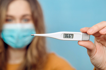 Close-up portrait of a girl in a medical mask on her face with suspected coronavirus and arm with outstretched thermometer. 2020 coronavirus epidemic. Covid-2019. Woman on a blue background.