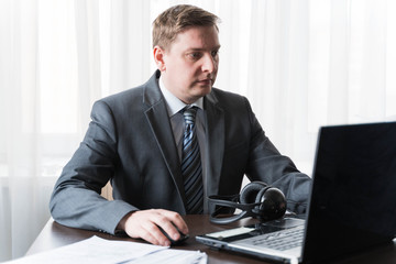 Young man in gray suit, shirt at telework due to quarantine of coronovirus pandemic COVID-2019.Remote work at home table.Communication on headset with boss,colleagues through laptop.Online conference