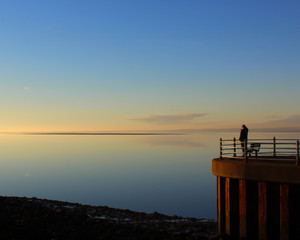 A lone figure stands looking out over the calm sea on a golden sunset at Morecambe in Lancashire