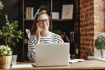 Young freelancer speaking on phone and works on a laptop in her home office