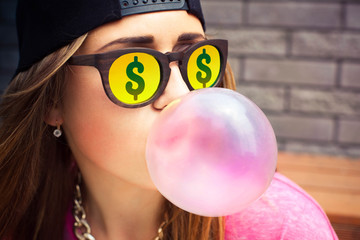 Young woman in sunglasses with dollar makes a chewing gum bubble