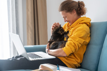 Happy young woman in a yellow sweatshirt works at home on a blue sofa with a laptop and a cat,...