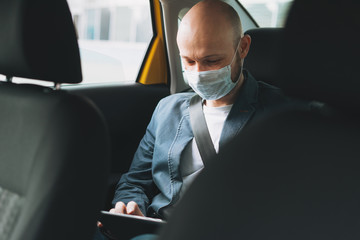 Bald man businessman in medical face mask using tablet mobile phone inside yellow car taxi, concept...