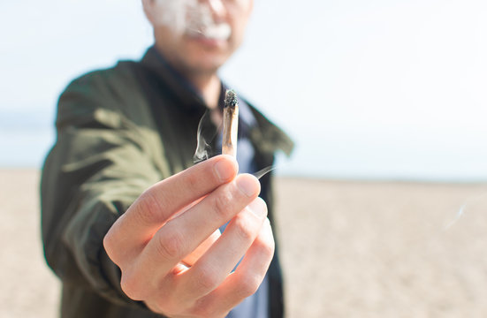Young man holding a lit marijuana joint while smoking on the beach. Blur background and copy space right.