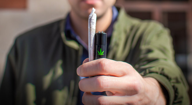 Young man on the street showing a big cannabis joint and a lighter with a marijuana leaf, close up. Blur background.