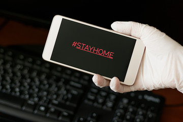 Hashtag stay home! The inscription on the smartphone screen. Female hand in a white glove holds a phone. The recommendation is quarantined at home to prevent coronovirus covid 19.