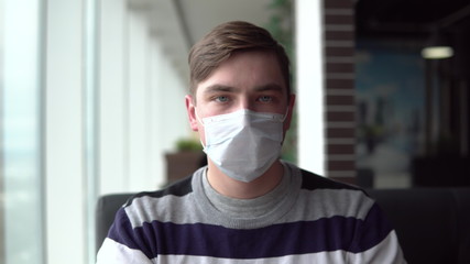 Young man in a medical mask. A man sits in a cafe by the window with a medical mask on his face. Prevention COVID-19.