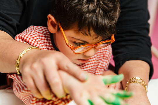 profesora educaci&oacute;n especial y alumno con bata est&aacute;n pintando con las manos una corona de cumplea&ntilde;os y una cartulina amarilla con pinturas acrilicas sobre la mesa. terapia de manualidades pl&aacute;sticas.