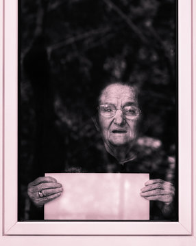 Old Sad Woman Showing Empty Signboard At The Window In Silent Request For Help - Concept Of Loneliness , Isolation And Social Unease For Elderly People - Dramatic Black And White Filter Look Image