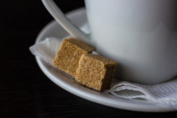 Two pieces of cane sugar on a saucer near white cup. Close up of brown pieces of sugar