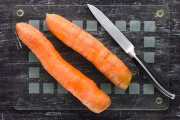 Top view of two unpeeled carrots and knife on glass cutting board before peeling on the black background