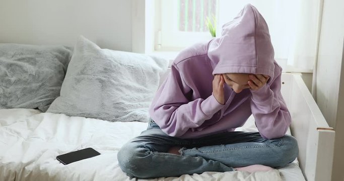 Depressed teenage girl sitting alone at home with smart phone. Upset adolescent teenager outcast wearing hood feeling worried or scared about social media cyber bullying mobile message concept.